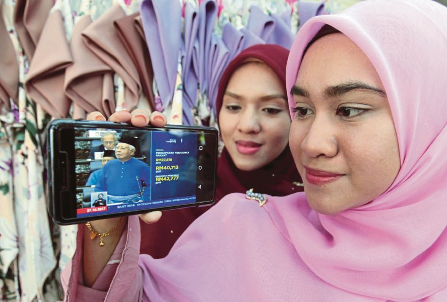 Peniaga dalam talian, Nurul Athirah Razali (kanan) bersama kakaknya, Siti Nur Fahimah Razali menyaksikan pembentangan Bajet 2018 oleh Perdana Menteri, Datuk Seri Najib Razak   melalui telefon pintar.